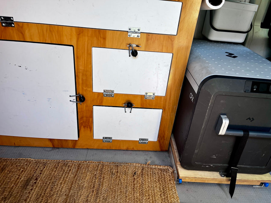 An rv rental's interior: custom wood cabinetry with white doors, a large Dometic CFX cooler, and a woven mat.