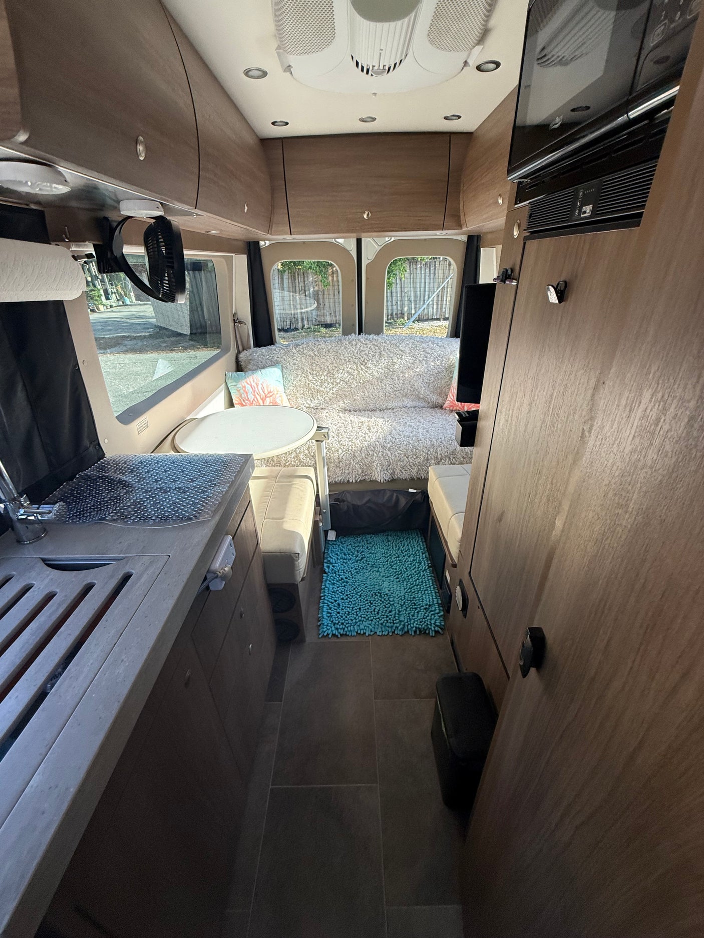 Inside a bright RV rental: compact kitchen, dining table, and a cozy lounge area with a fluffy white bed at the rear.