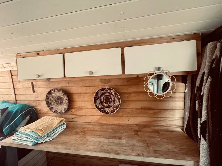 Inside an RV rental: cozy wooden interior with white cabinets, a desk, woven wall art, and stacked towels. — photo 13