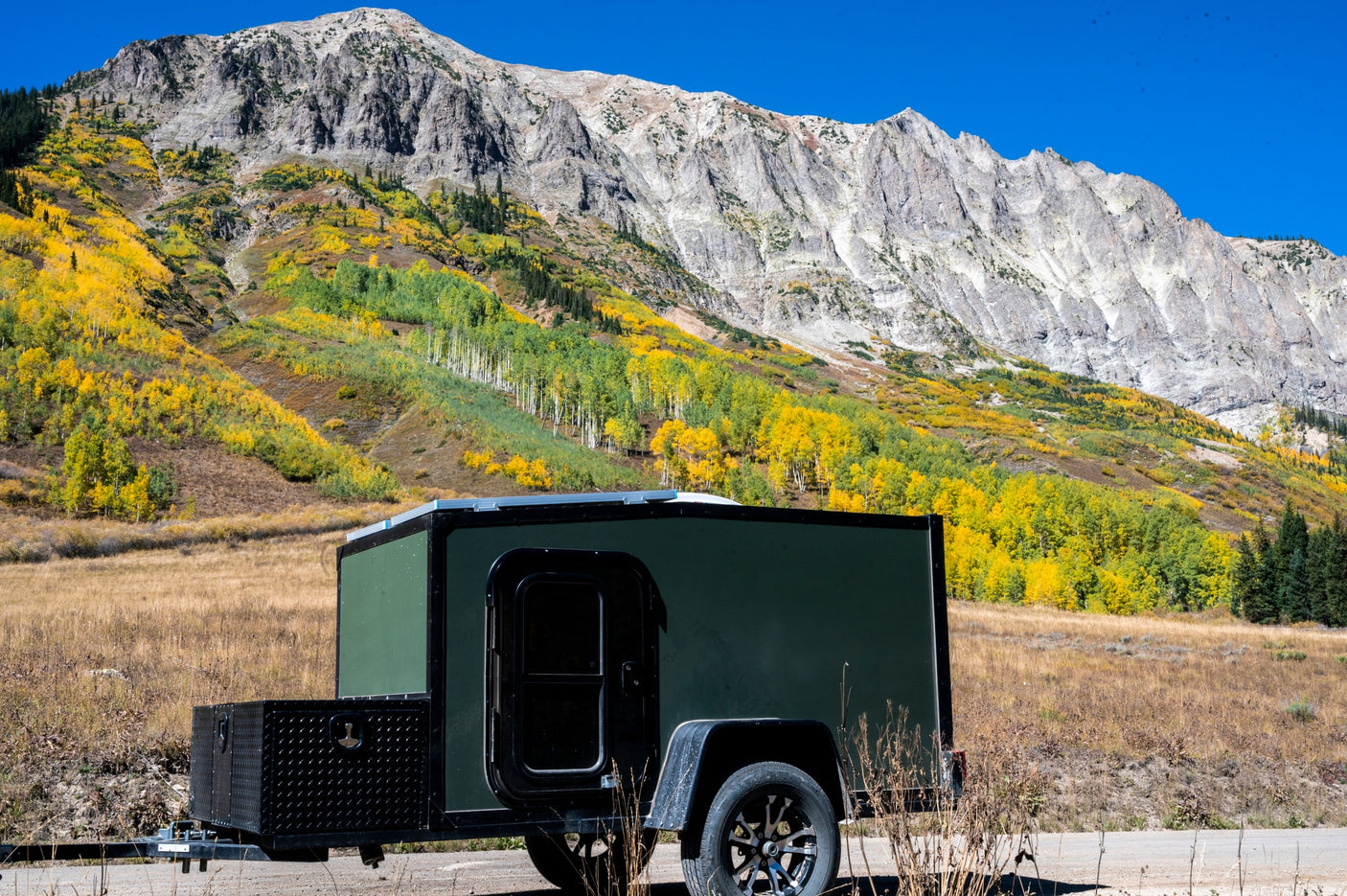 Dark green teardrop RV rental camper parked in autumn mountains, surrounded by vibrant yellow and green aspen trees under a blue sky.
