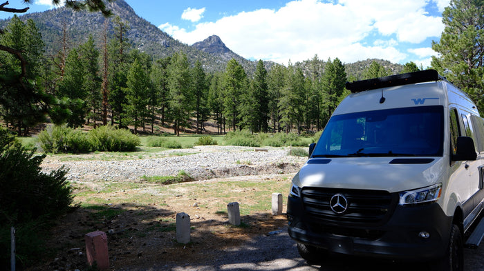 parked among mountains and pine trees under blue sky.|Enjoy an RV rental adventure! Two people camp by their Winnebago camper van and campfire in a scenic outdoor setting.|Explore nature with an RV rental! A white Mercedes Sprinter camper van is parked among pine trees