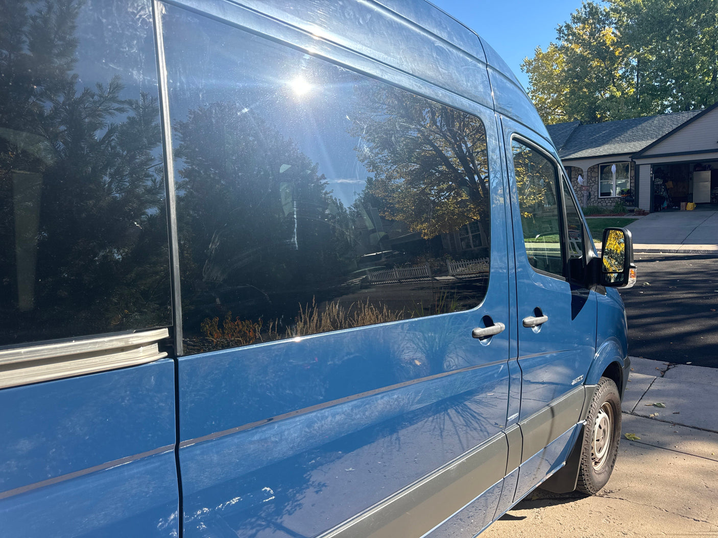 Blue Sprinter van for RV rental, parked under sun, reflecting trees and a house on its windows.