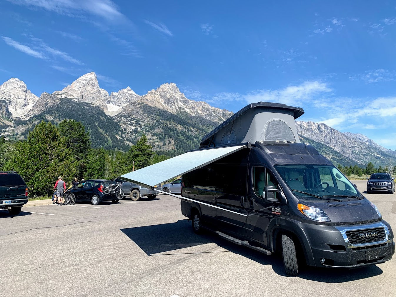 Dark gray RV rental with pop-top & awning deployed, parked in a lot with majestic Teton mountains & clear sky. Ready for adventure.
