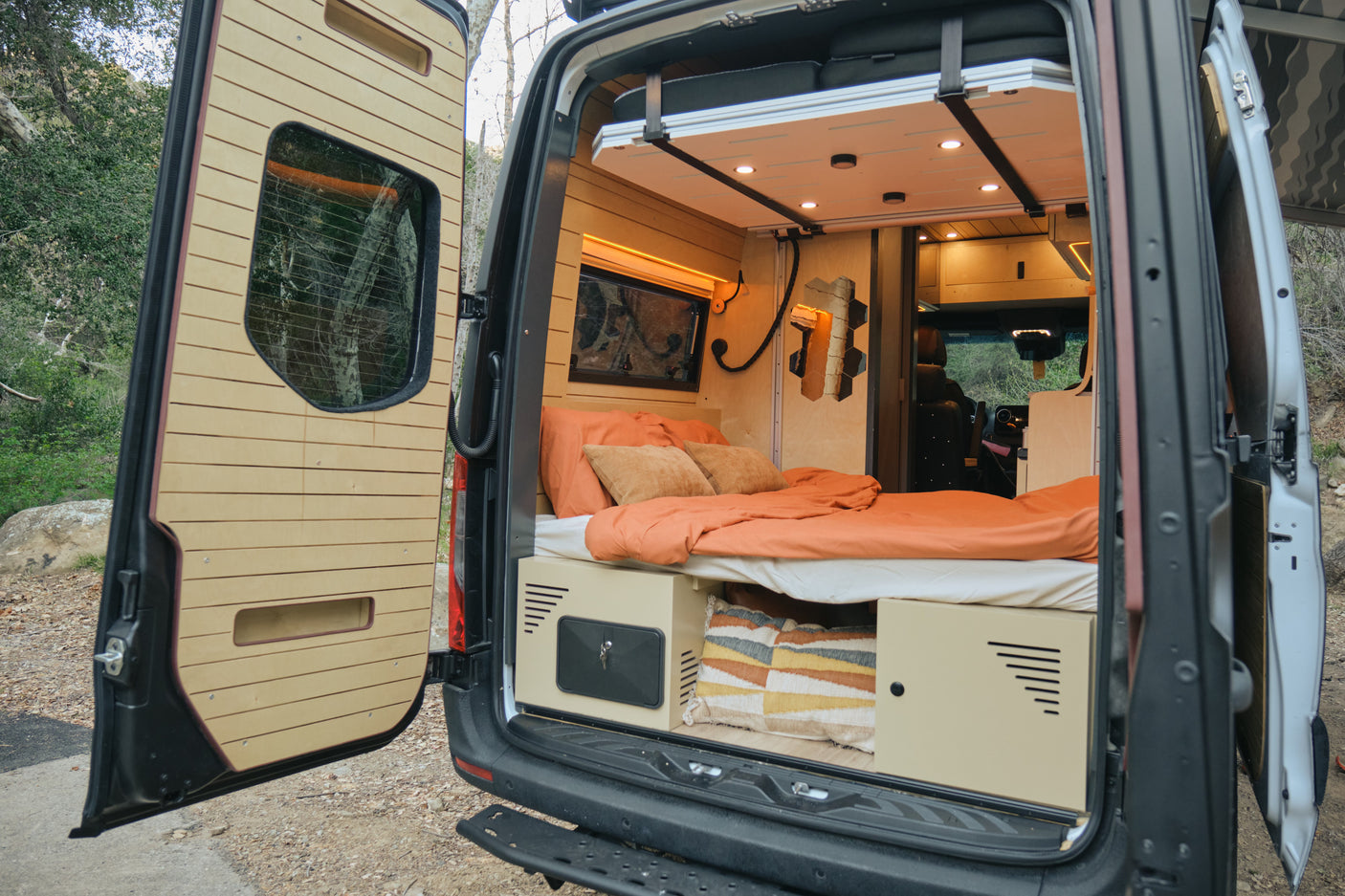 Cozy camper van interior of an RV rental, featuring a bed, storage, and custom wooden walls, open to nature.