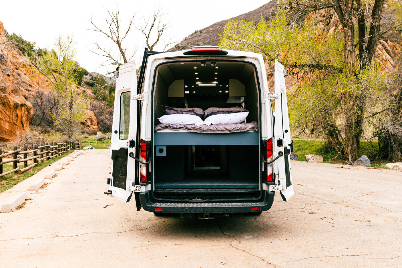 Rear view of a white camper van with doors open, revealing a comfy bed and storage.