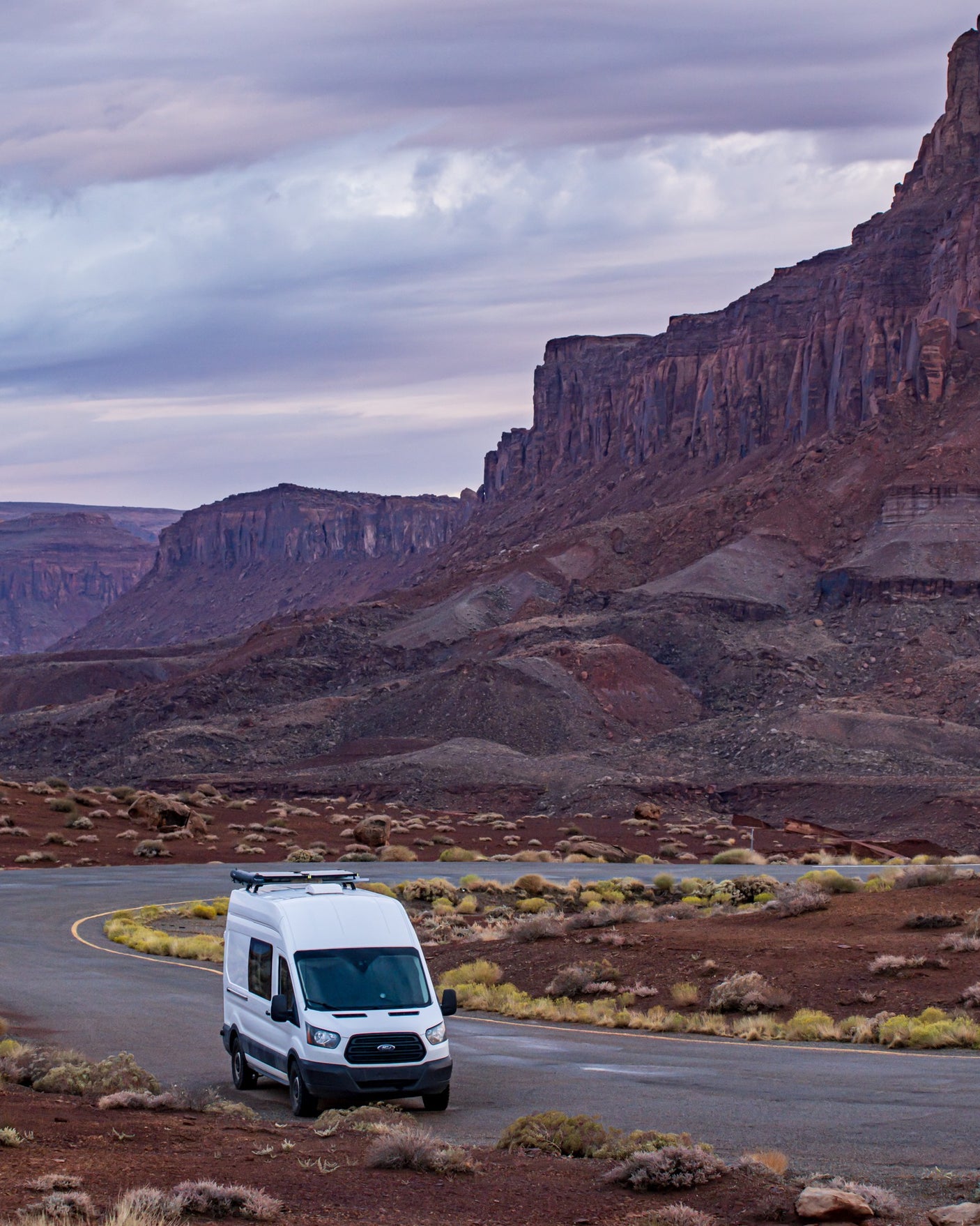 White rv rental campervan parked on a winding desert road, surrounded by majestic red cliffs and a dramatic sky.