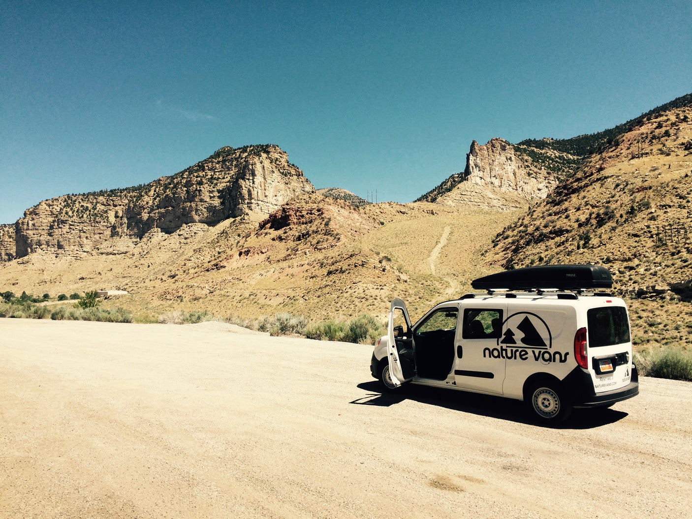 Explore the wild! White Nature Vans camper van, an RV rental option, parked on a dirt road in arid mountains under blue sky.