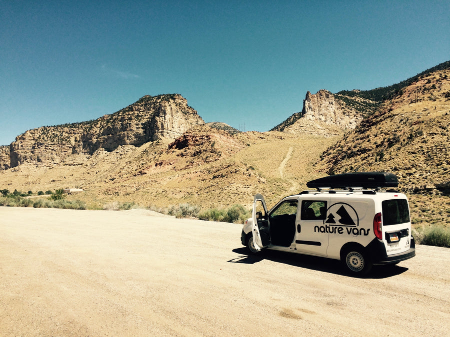Explore the wild! White Nature Vans camper van, an RV rental option, parked on a dirt road in arid mountains under blue sky.