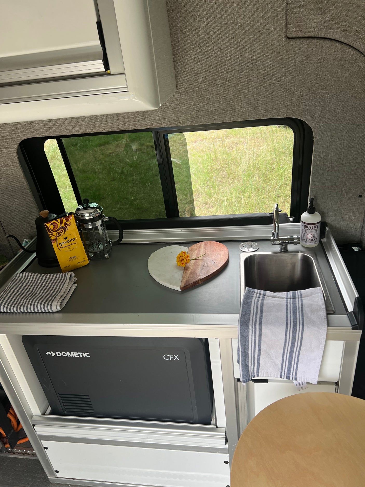 Compact RV rental kitchen with sink, Dometic fridge, and coffee setup. Enjoying van life with a view of green fields.