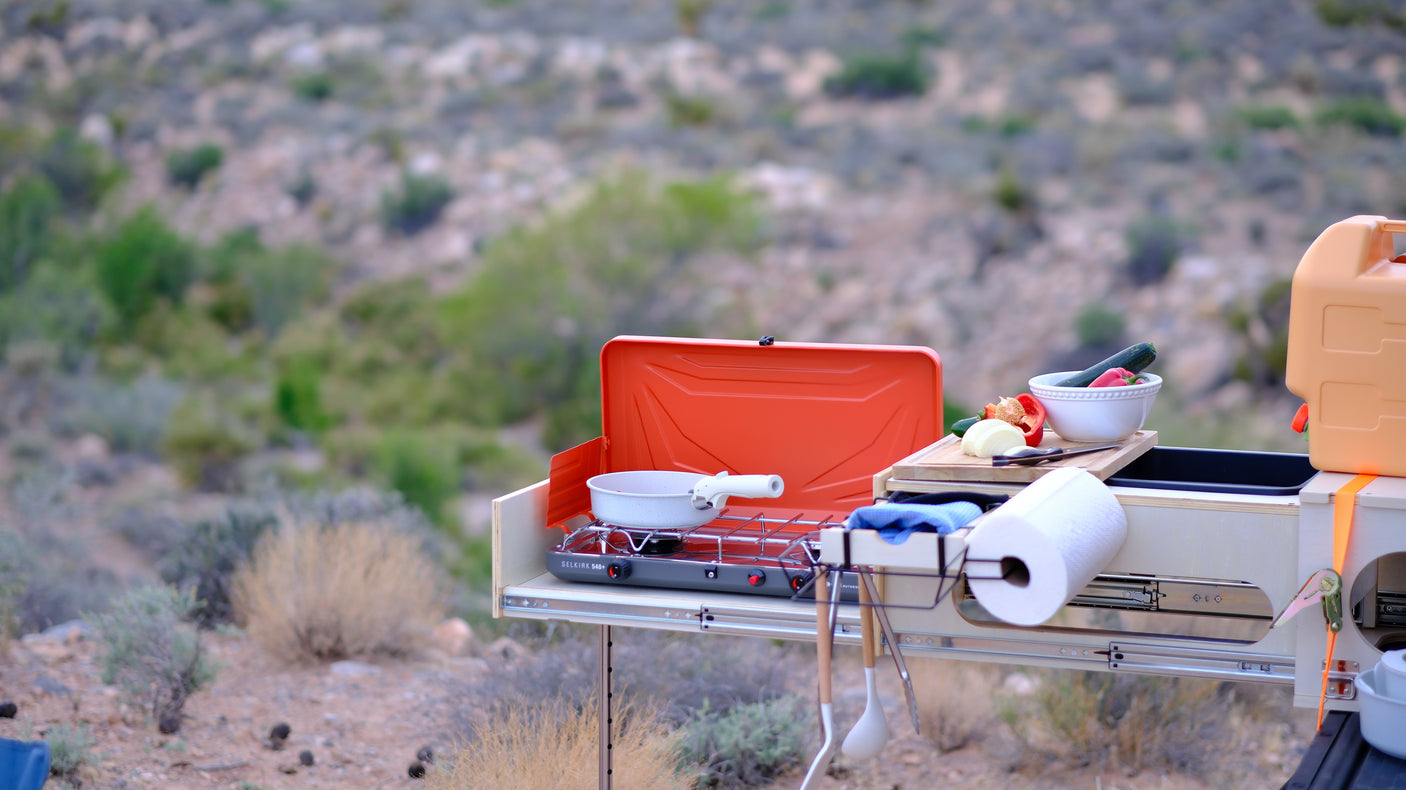 RV rental portable kitchen setup featuring a camping stove, pan, fresh veggies, and utensils, ready for camp meals in a desert setting.