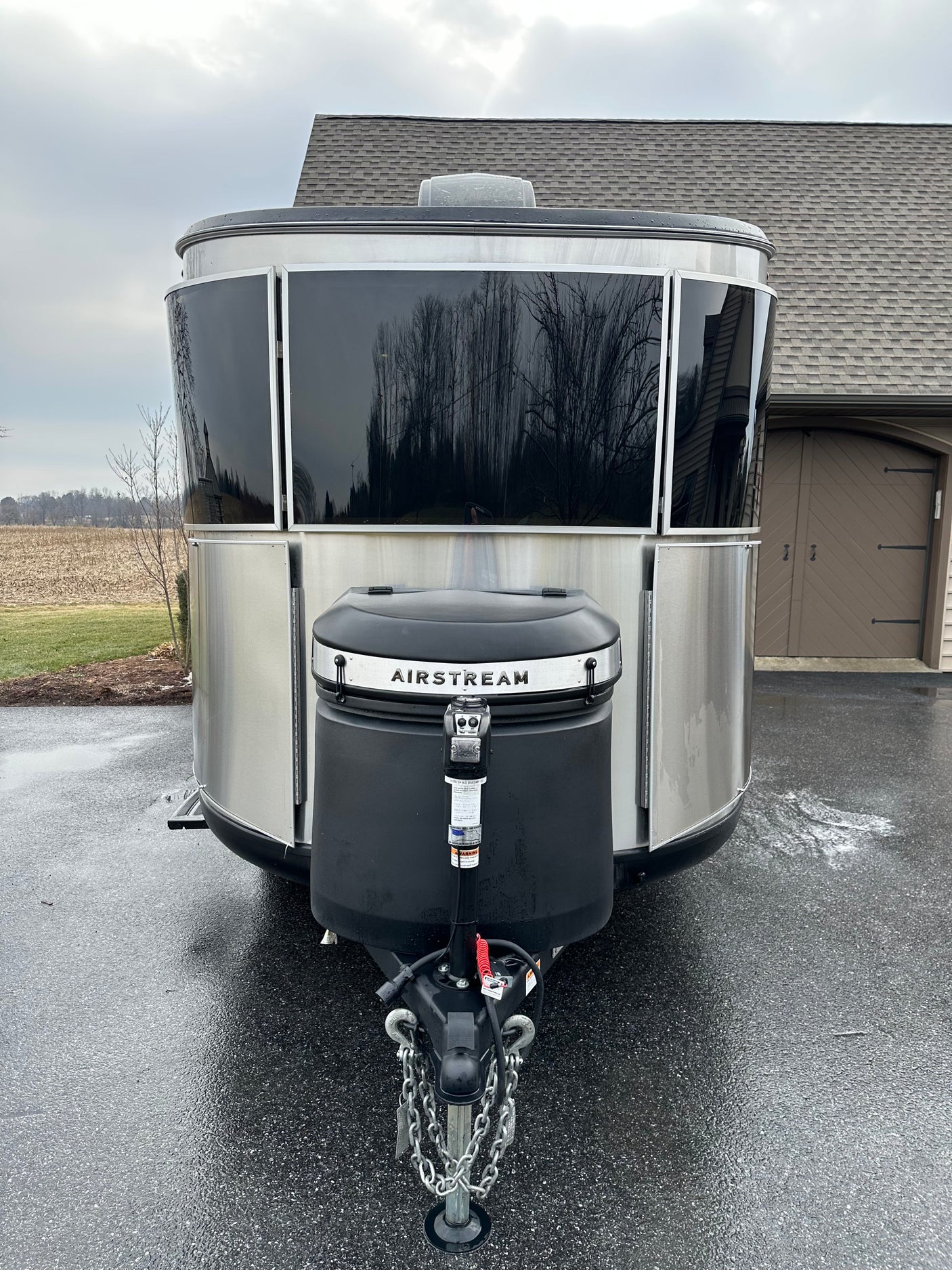 A sleek silver Airstream travel trailer, ideal for an RV rental. Parked on a wet driveway, reflecting trees and a house.