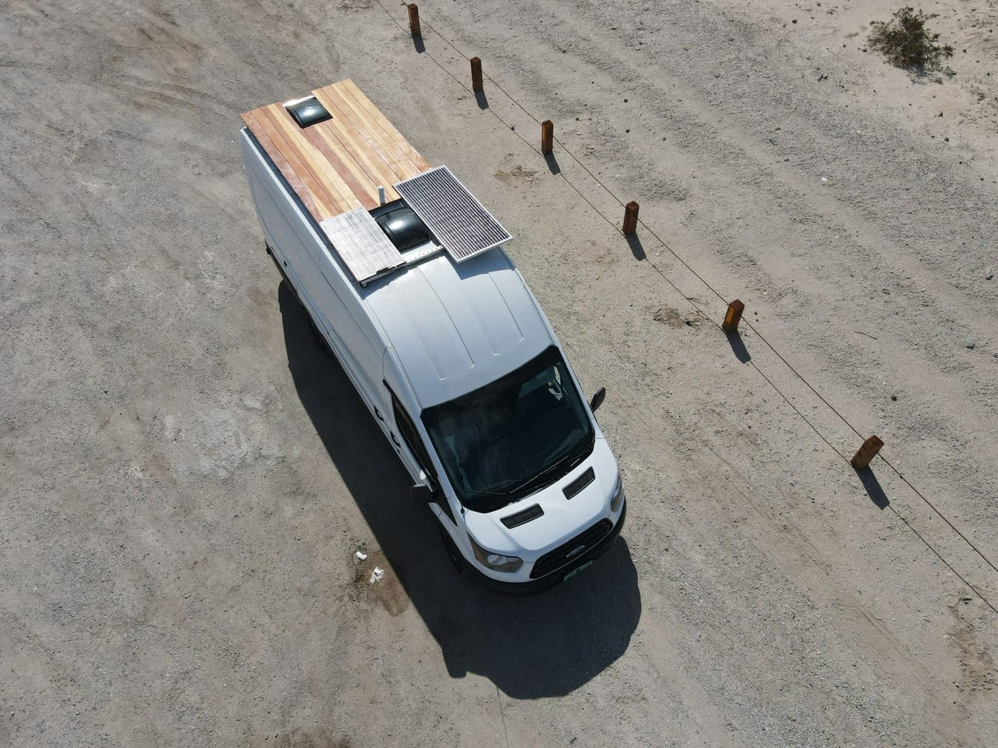 RV rental: Aerial view of a white camper van with a wooden rooftop deck, solar panels, and skylight, parked on a gravel road.