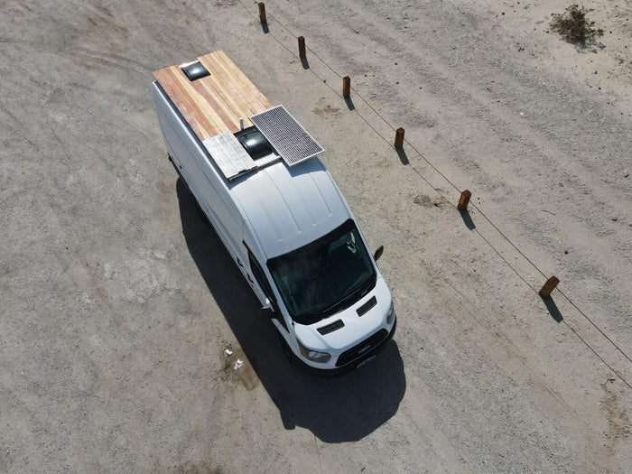 RV rental: Aerial view of a white camper van with a wooden rooftop deck, solar panels, and skylight, parked on a gravel road.