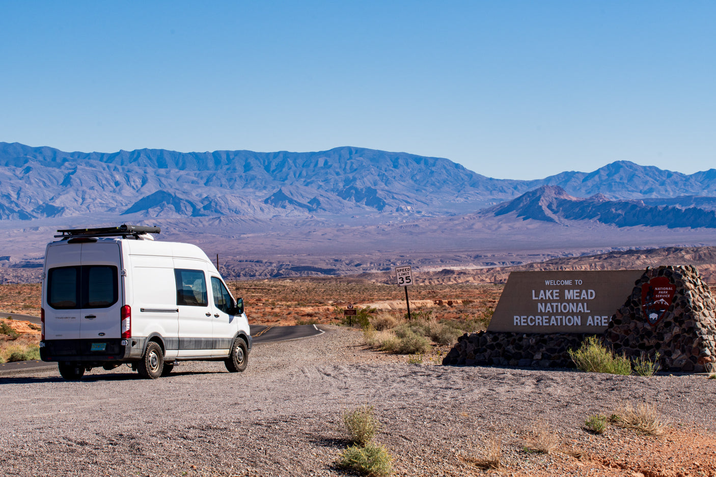 An RV rental van pauses at the entrance to Lake Mead National Recreation Area, with vast desert mountains behind.