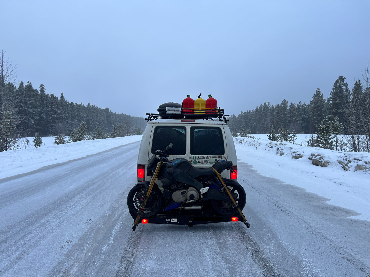 Winter RV rental adventure: White van with motorcycle on snowy road, surrounded by snow-covered pines.
