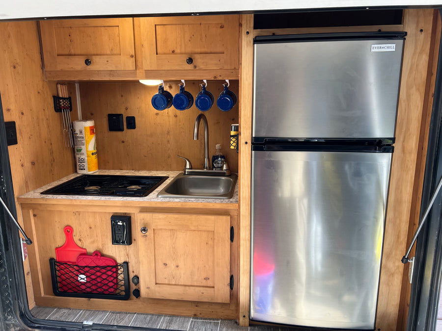 Inside an RV rental, a compact kitchen features wood cabinets, a stainless steel fridge, stove, sink, and essential accessories.