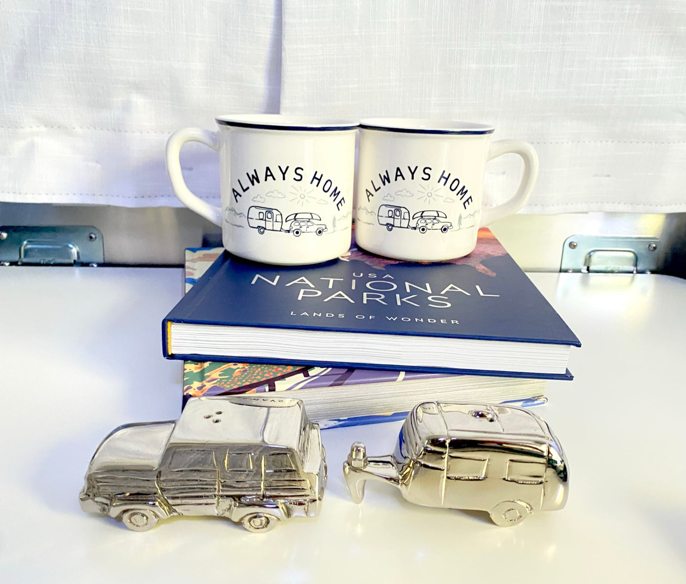 Travel-themed decor: 'Always Home' mugs with car/RV graphics, a National Parks book, and silver car/RV shakers. Great for rv rental enthusiasts.