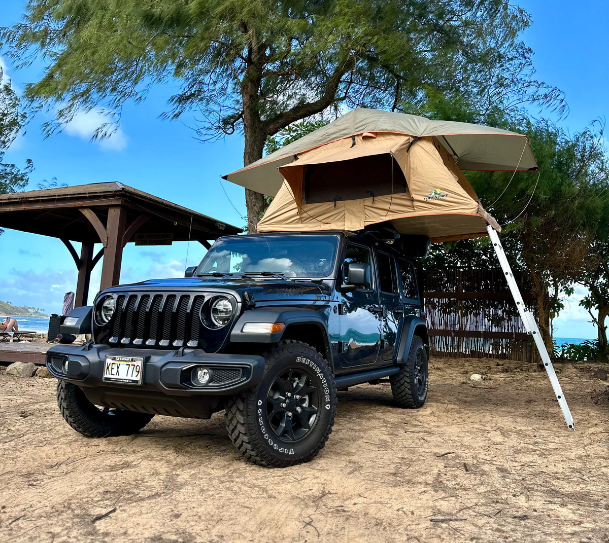 Rooftop tent on a black Jeep Wrangler parked on sand near a beach and gazebo under a tree. Great **rv rental** for adventurous trips. — photo 2