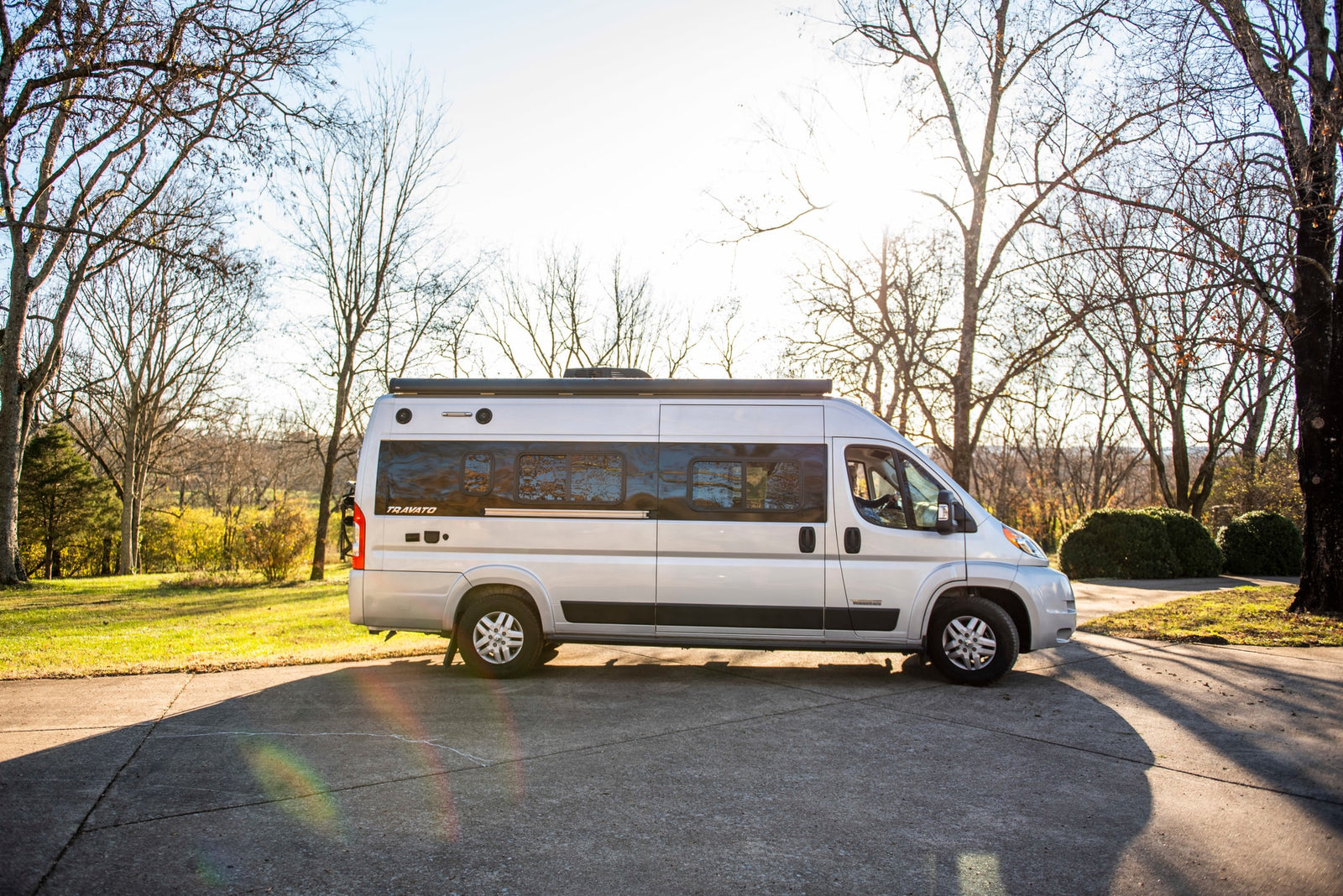 Silver Winnebago Travato camper van rv rental, parked on a sunlit pavement amidst bare trees and green grass.