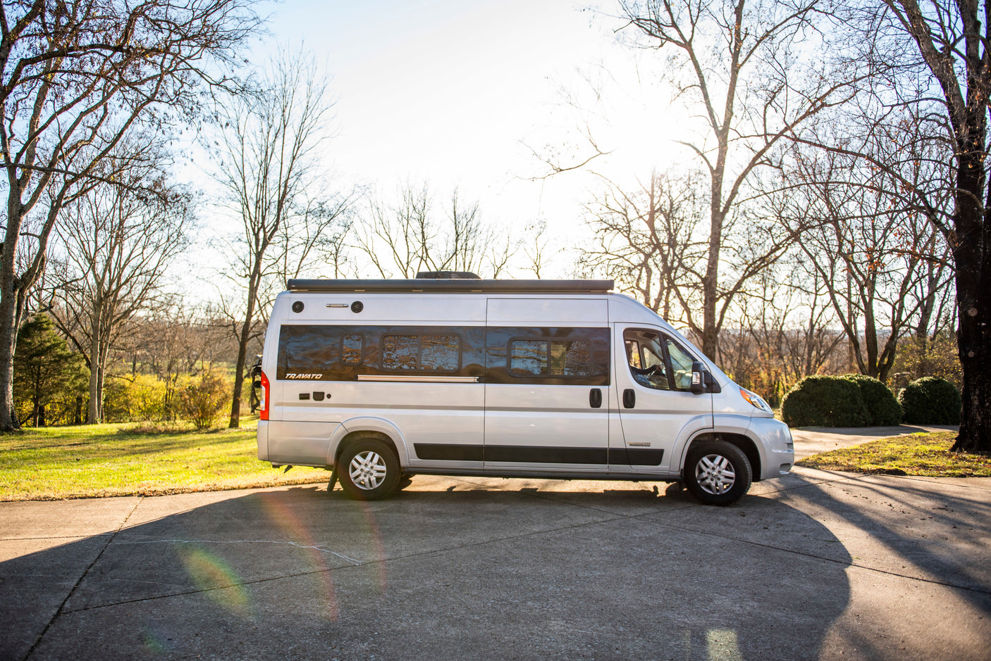 Silver Winnebago Travato camper van rv rental, parked on a sunlit pavement amidst bare trees and green grass.