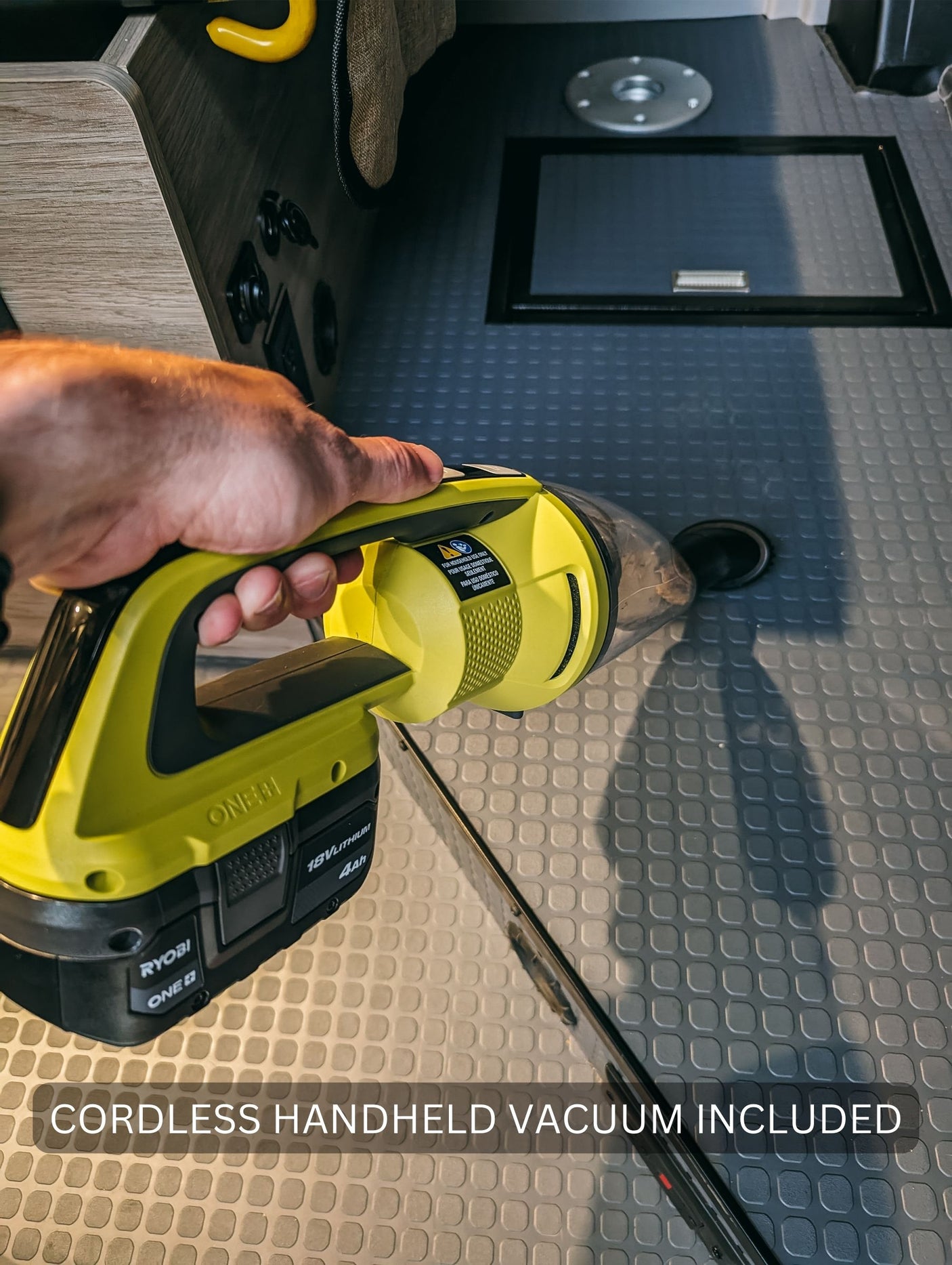 Inside an RV rental, a hand uses a Ryobi cordless handheld vacuum on the gray textured floor. Cleaning convenience included for trips.