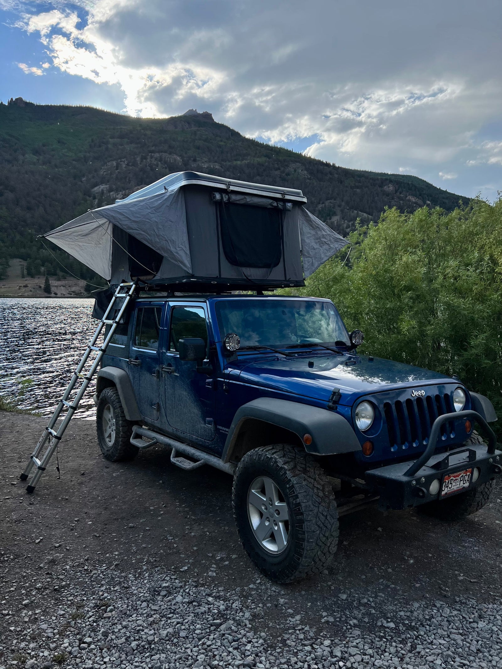 This blue Jeep with a deployed rooftop tent by a lake offers an amazing RV rental experience amidst scenic mountains.