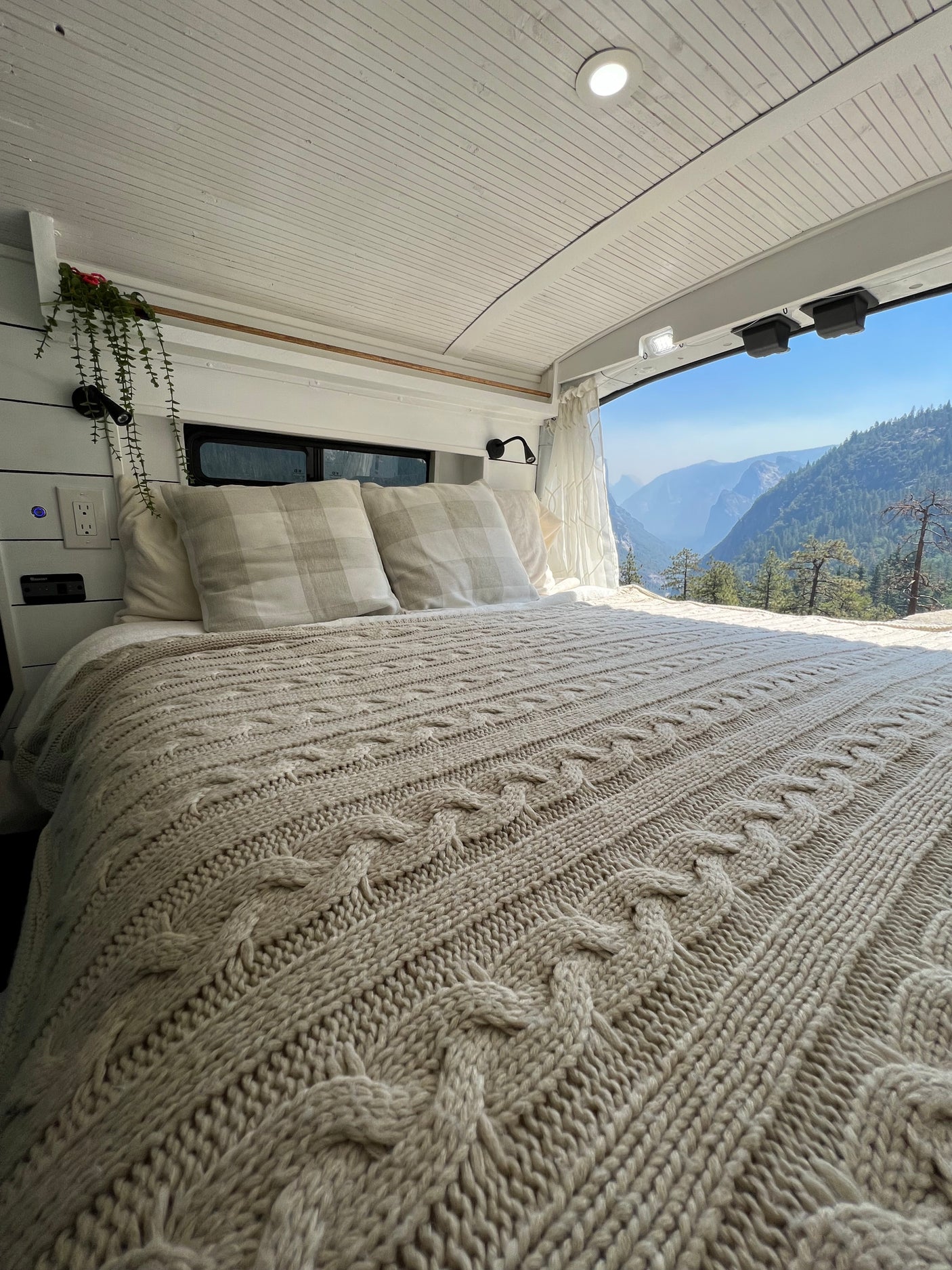 Cozy RV rental bed with a cable knit blanket and mountain view. Perfect for van life adventures, offering comfort in nature.