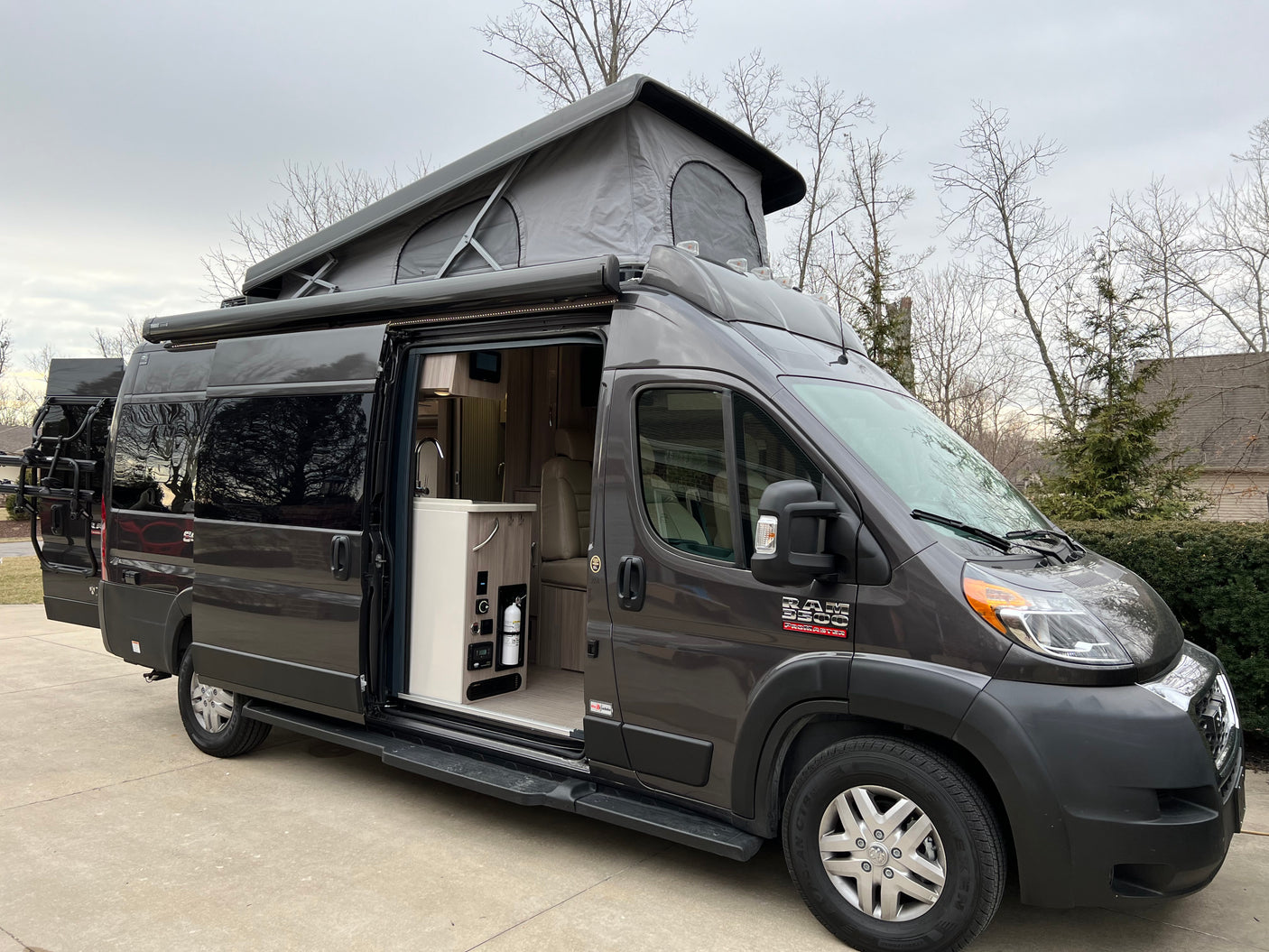 Experience adventure! This modern grey pop-top camper van RV rental features an open side door, spacious interior, and rear bike rack.