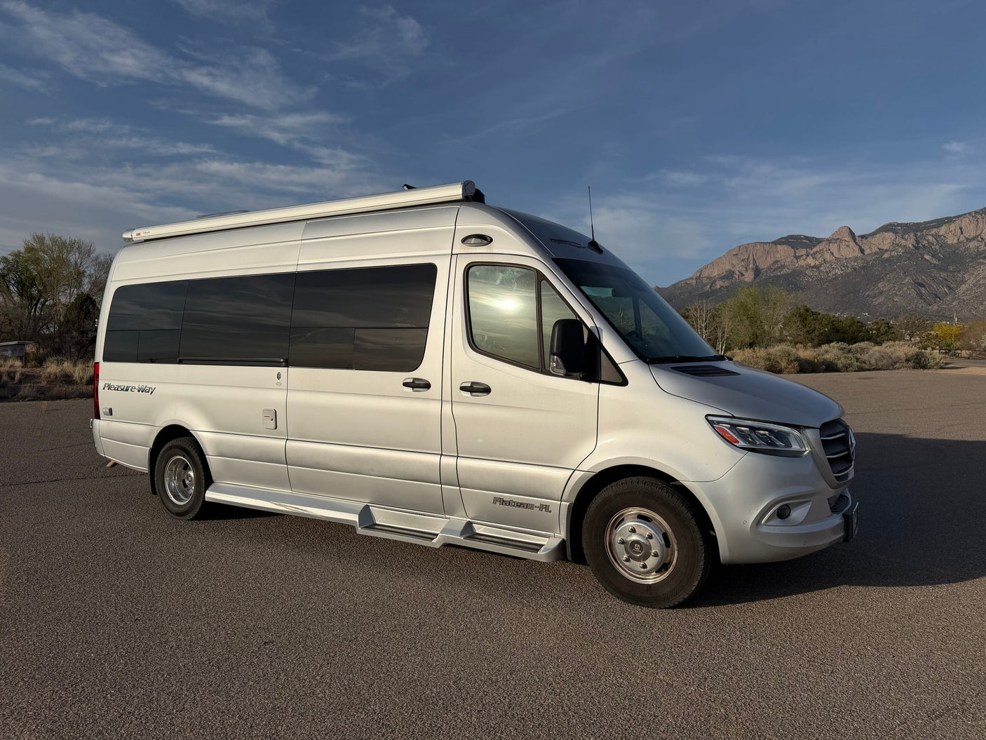 Silver Pleasure-Way rv rental van parked on asphalt, mountains and blue sky in background.