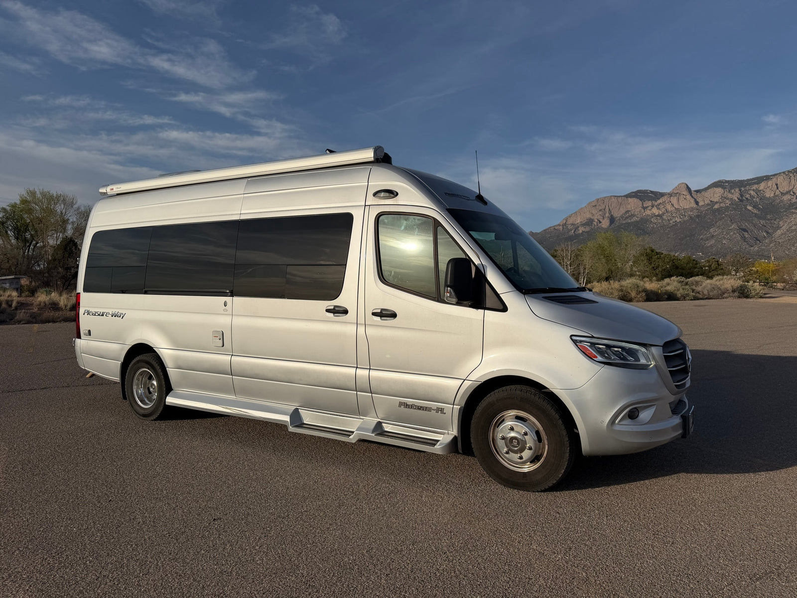 Silver Pleasure-Way rv rental van parked on asphalt, mountains and blue sky in background.