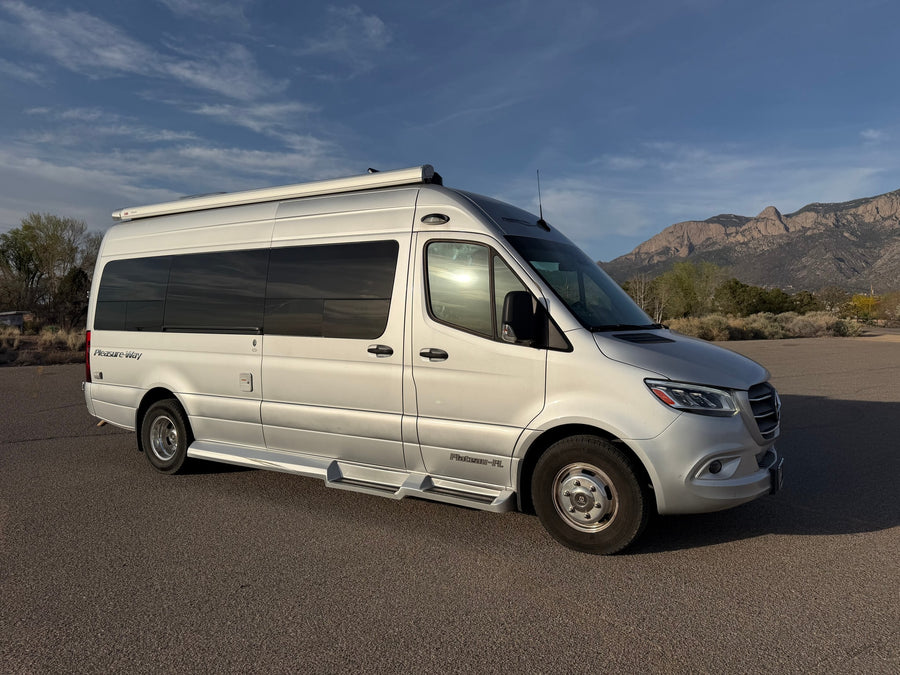 Silver Pleasure-Way rv rental van parked on asphalt, mountains and blue sky in background.