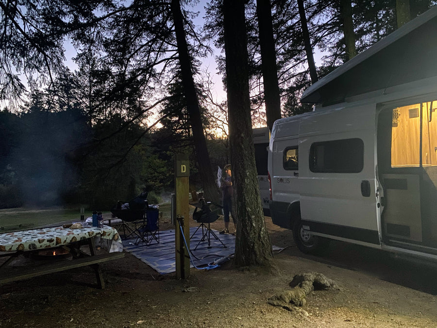 Enjoying a serene evening at a forest campsite with a Winnebago Solis RV rental, campfire glowing, and person busy.