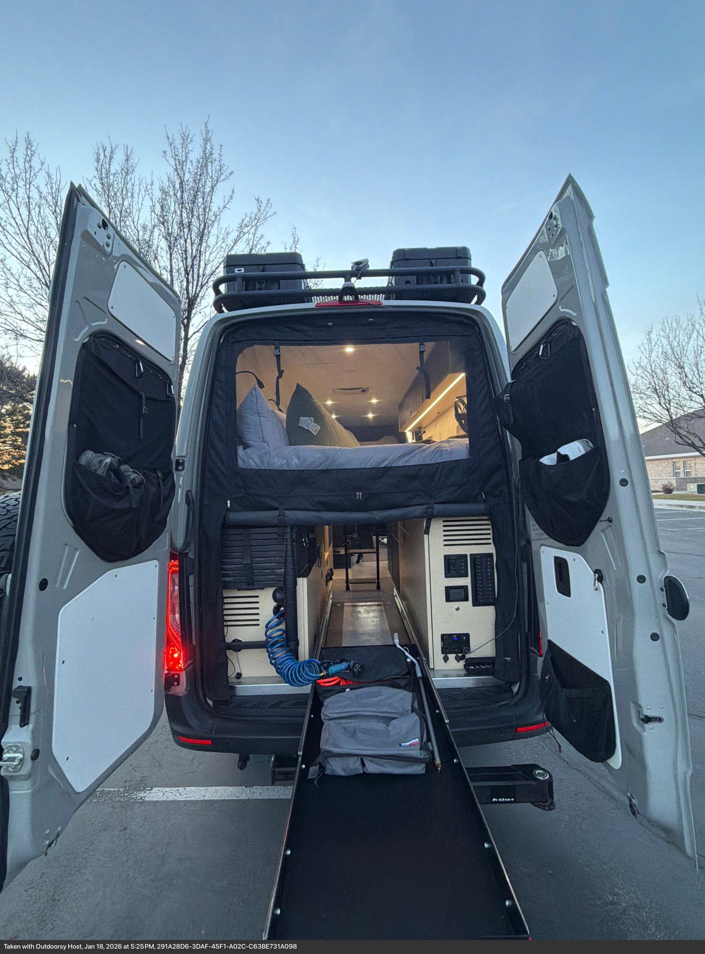 RV rental: Back of a camper van with open doors, showing a cozy bed, interior lighting, and a pull-out ramp for gear.