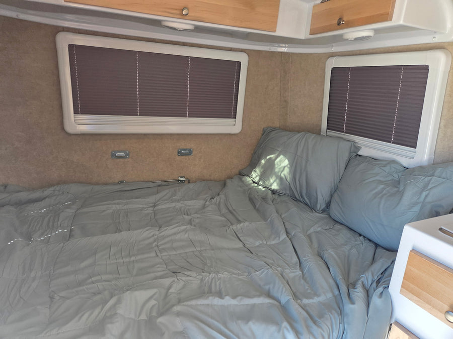 Cozy RV rental interior with a light grey bed, pillows, and windows with blinds, perfect for comfortable travel.