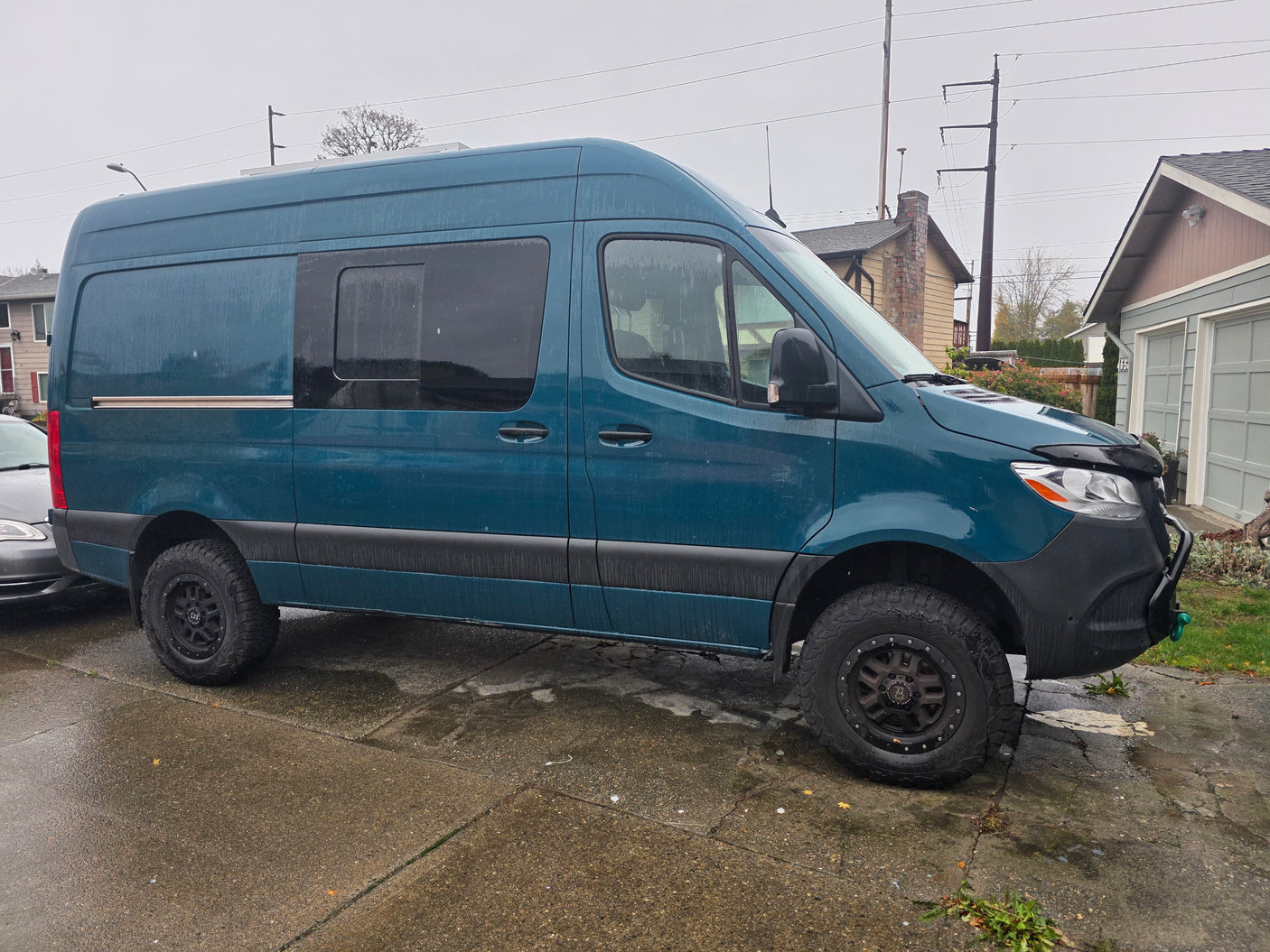 Teal adventure van, an ideal RV rental, parked on a wet drive. Features include off-road tires and roof solar panels.