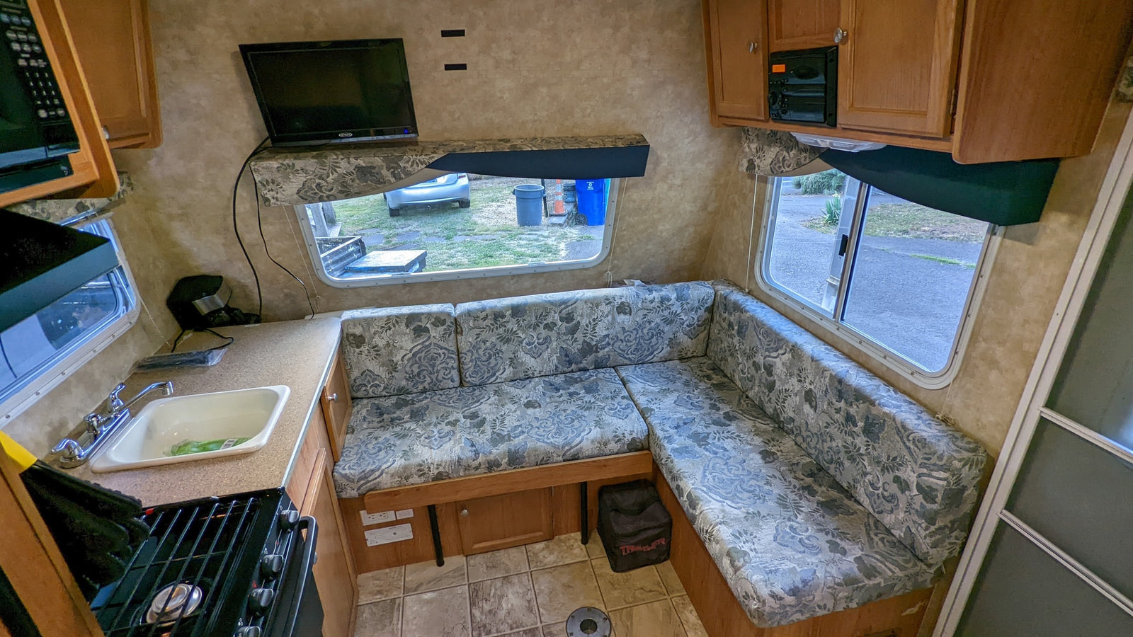 Inside an RV rental: a compact kitchen, L-shaped dinette with floral cushions, and mounted TV. Tiled floor and wood cabinets.