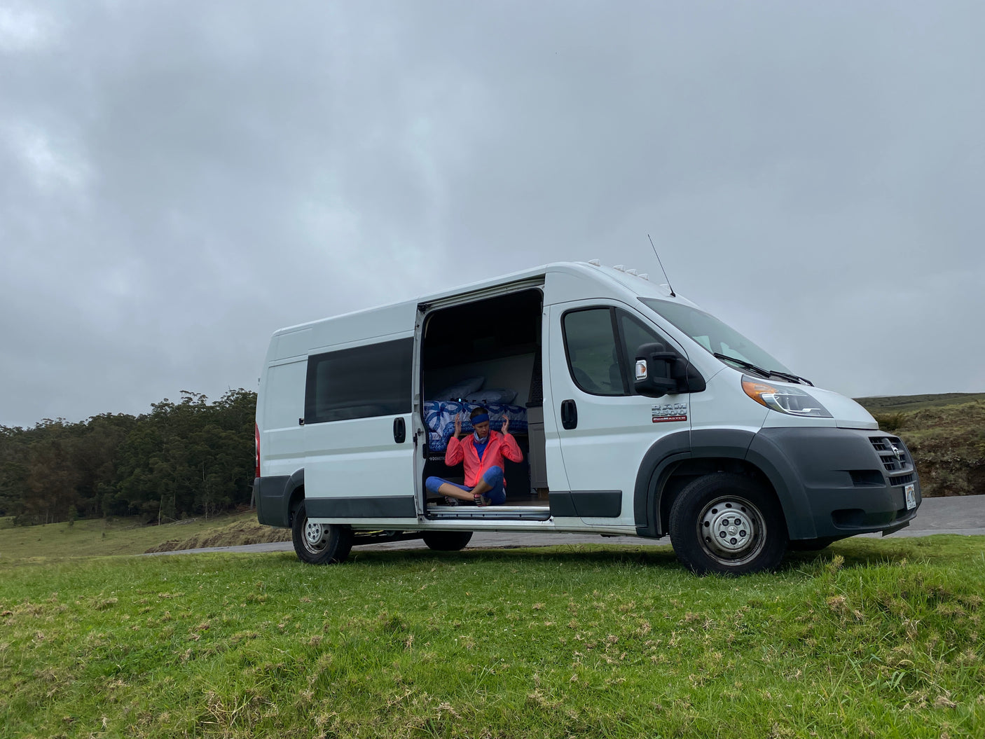Person in a serene pose inside a white RV rental van, parked on a grassy hill beside a road under an overcast sky.