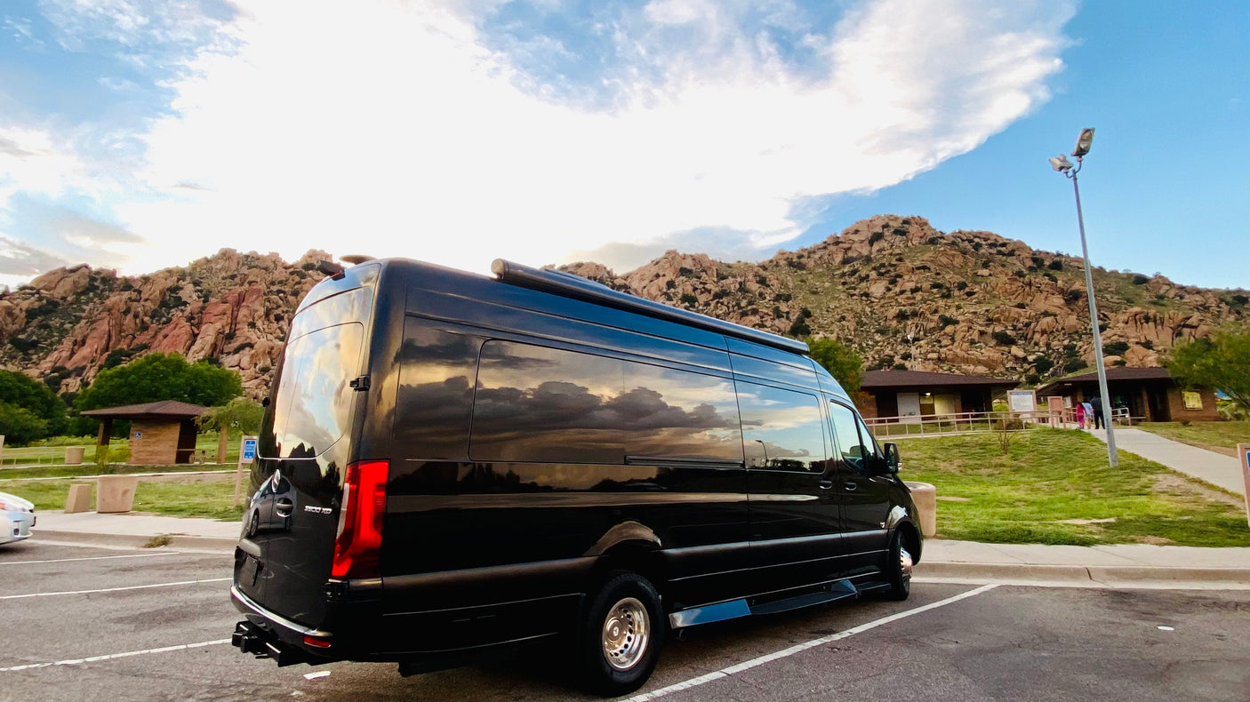 A black Mercedes Sprinter RV rental parked at a scenic mountain rest stop, reflecting clouds and sky on its shiny surface.