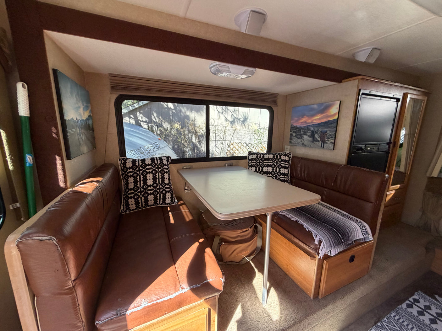 Cozy RV rental dining area: brown faux leather benches, table, window with outdoor view, and wall art create a welcoming travel space.