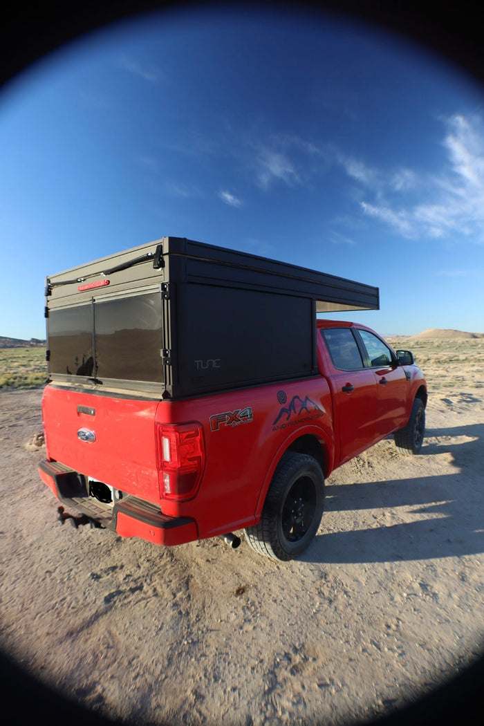 ideal for an RV rental or overland trip. Parked in a dusty desert landscape under blue skies.|Red Ford Ranger pickup truck with a Tune camper