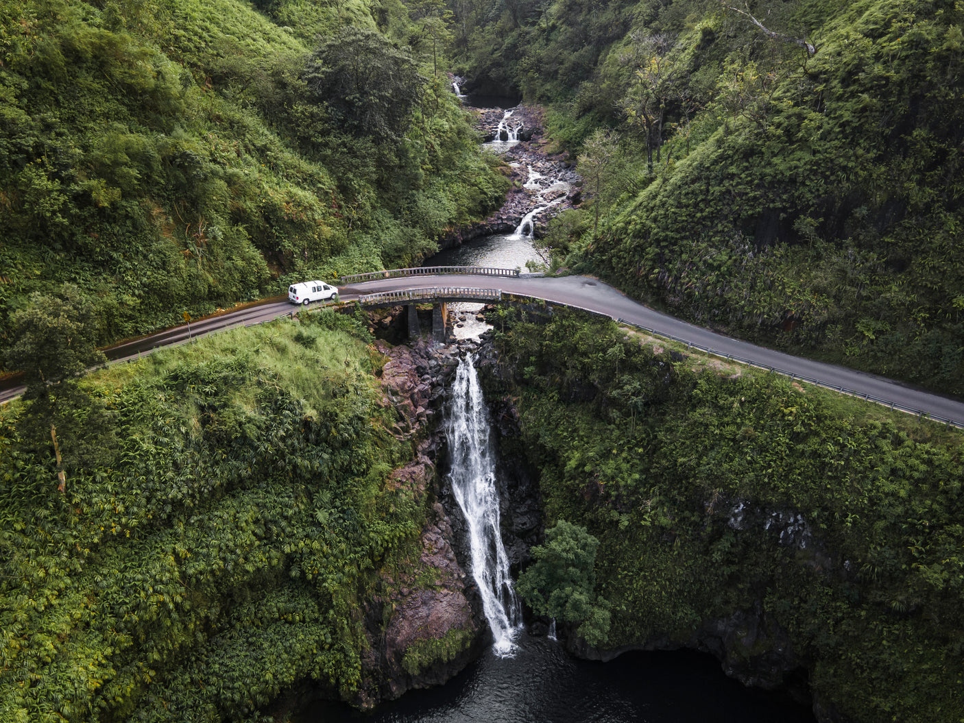 Scenic road trip via RV rental: A white van crosses a bridge above a stunning waterfall amid lush green valleys and smaller falls.