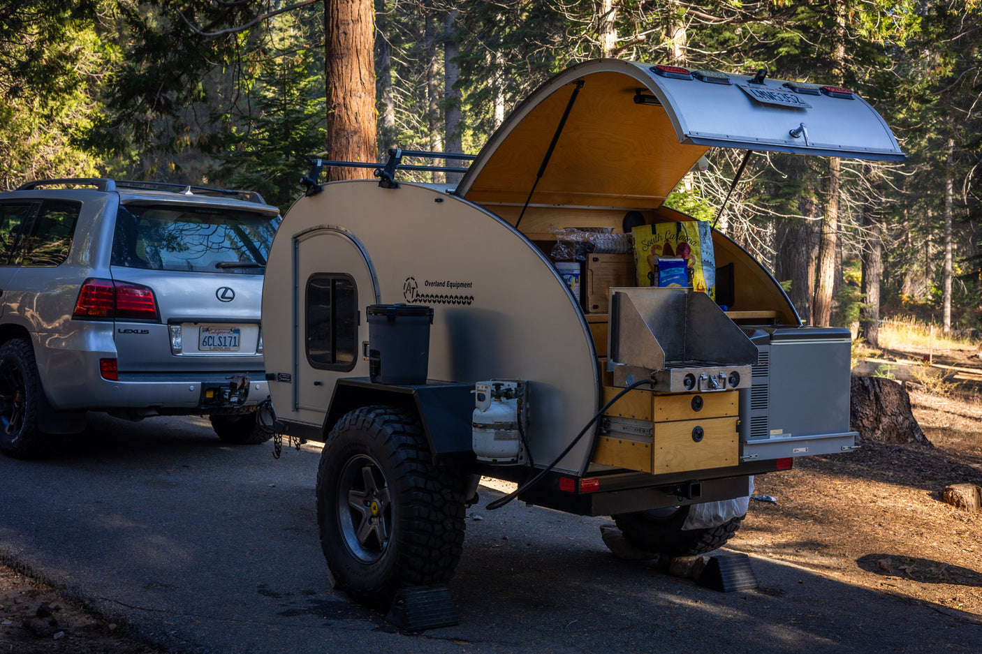 Overland Equipment teardrop RV rental setup with outdoor kitchen, ready for camping in a forest. Lexus SUV attached.