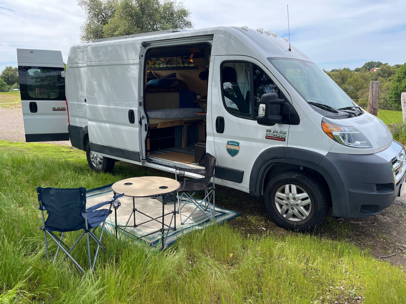 This white Ram ProMaster camper van, available for RV rental, offers an outdoor camping setup with chairs, table, and rug.