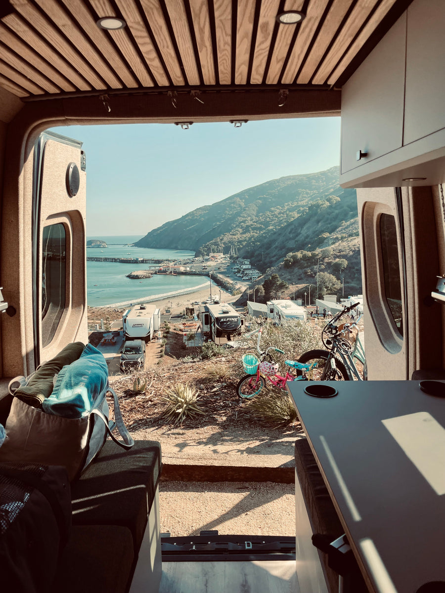 Inside an RV rental, a stunning view of a coastal campground, complete with beach, pier, and lush hills.