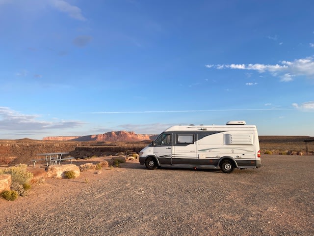 surrounded by mountains under a blue sky.|Scenic RV rental stop: A camper van overlooks red rock desert mesas under a vast blue sky.|Cozy RV rental interior featuring passenger seats