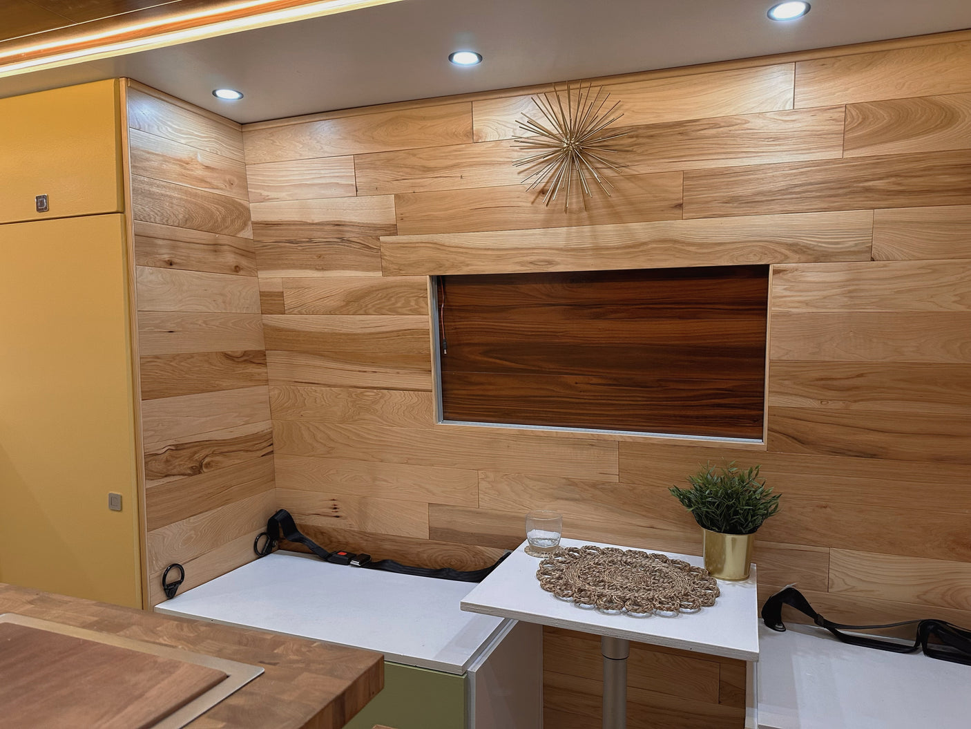 This RV rental interior showcases warm wood paneled walls, bright lighting, yellow cabinets, and compact seating with a table and plant.