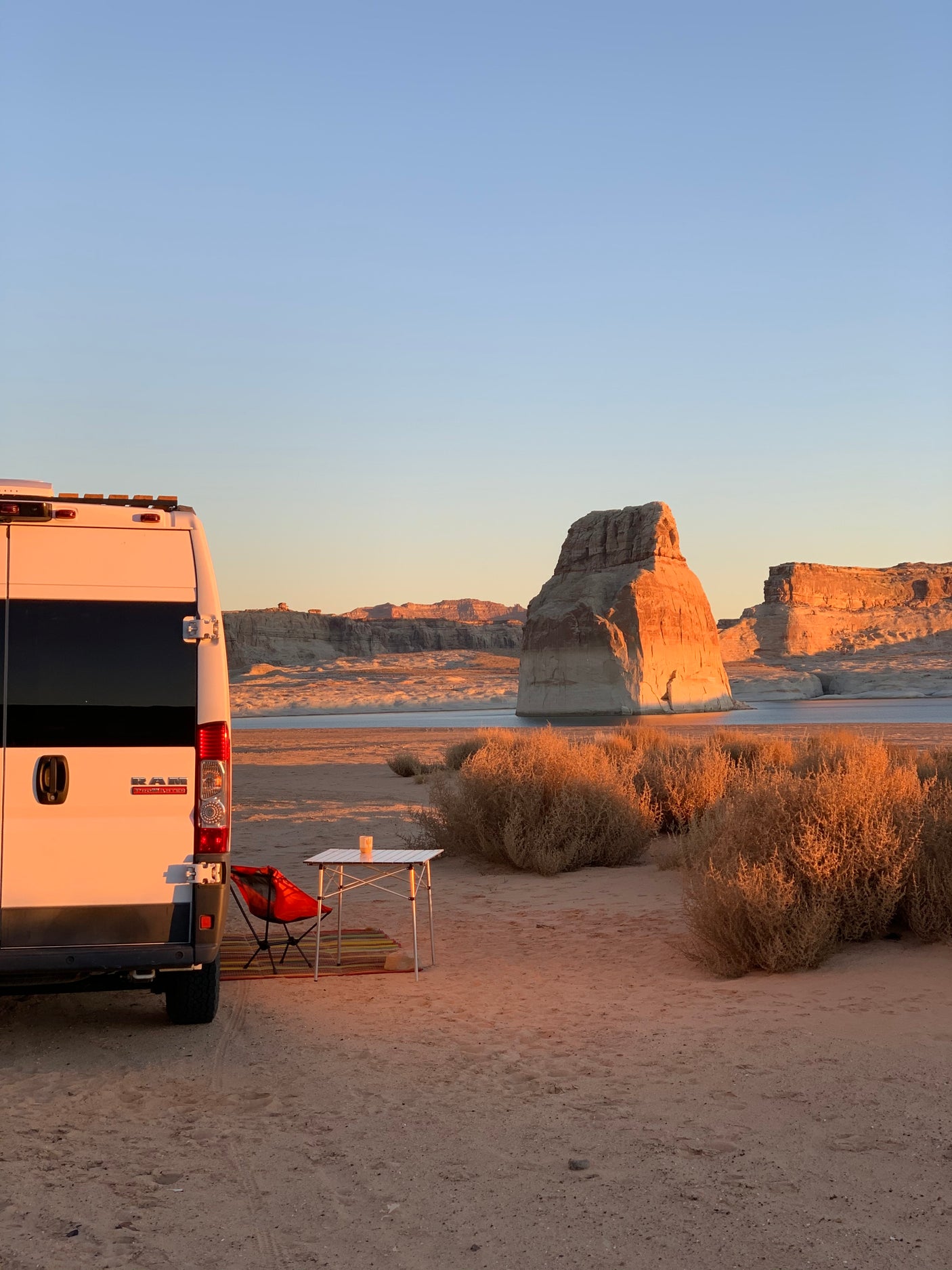 RV rental campsite at golden hour! A white camper van with a table & chair overlooks stunning desert rock formations & lake.
