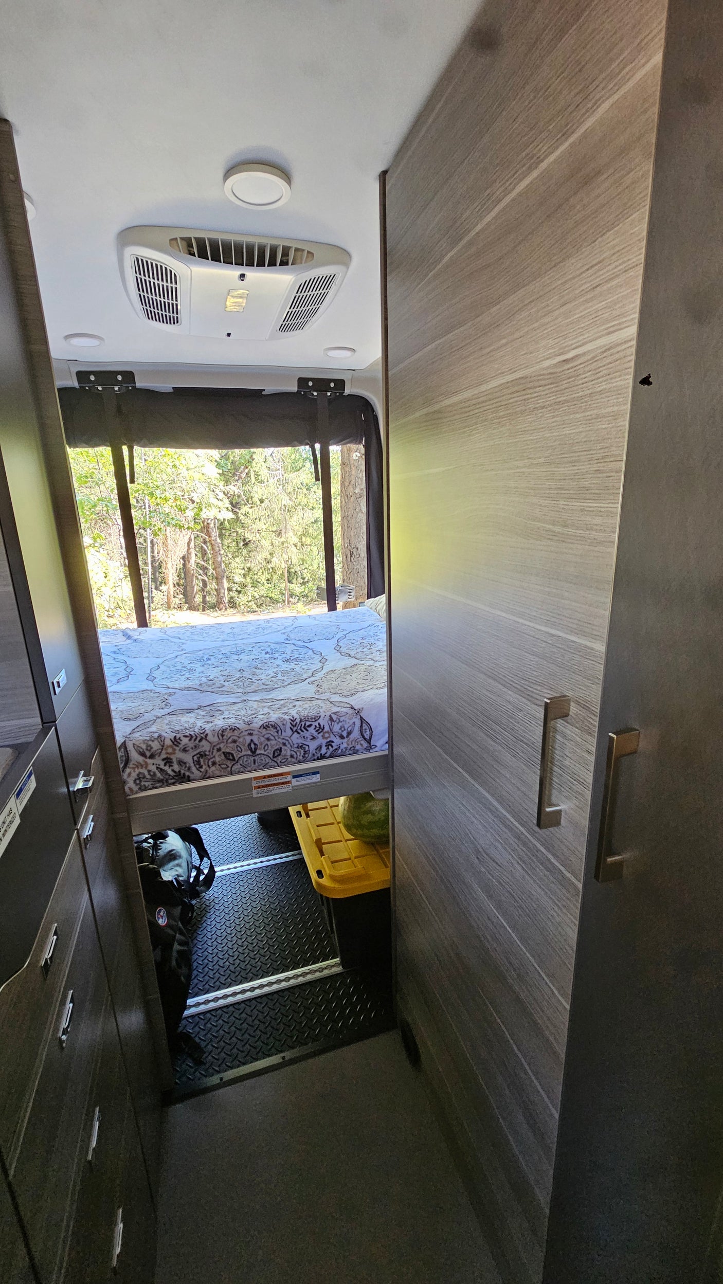 Inside an RV rental: rear bed, large window with forest view, AC, and under-bed storage. Ideal for travel.