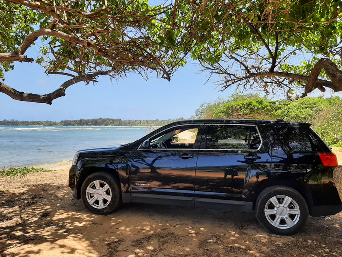A black GMC Terrain SUV is parked under a shady tree by a beautiful beach. Perfect for coastal adventures, like those found with an RV rental.