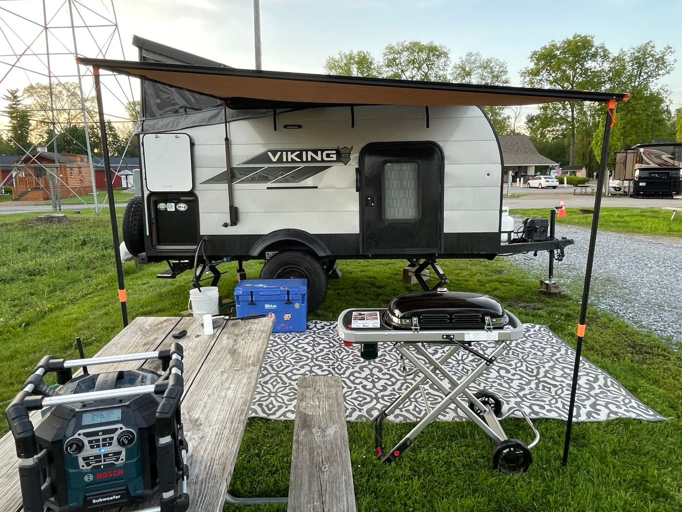 This RV rental showcases a compact Viking travel trailer with an extended awning, outdoor grill, and picnic table at a serene campground.